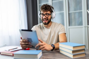 Gözlüklü genç bir öğrenci kitaplarla bir masada oturuyor, online bir derse katılıyor ve bir profesörle tabletinde video aracılığıyla konuşuyor, tamamen öğrenmeye odaklanıyor..