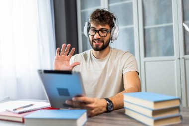 Gözlüklü genç bir öğrenci kitaplarla bir masada oturuyor, online bir derse katılıyor ve bir profesörle tabletinde video aracılığıyla konuşuyor, tamamen öğrenmeye odaklanıyor..