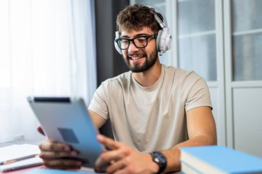 Gözlüklü genç bir öğrenci kitaplarla dolu bir masada oturuyor, tabletindeki sınava çalışmaya odaklanmış, notlarla çalışırken kendini adamış..