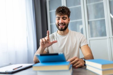 Genç bir öğrenci masada kitaplarla oturuyor, işaret dilini öğreniyor ve tabletinde el hareketleri yapıyor, iletişim becerilerini geliştirmeye odaklanıyor..