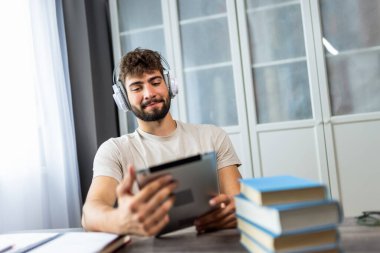 Kulaklık takan genç bir öğrenci kitaplarla dolu bir masada oturuyor, tabletindeki sınava çalışmaya odaklanmış, notlarla çalışırken kendini adadığını gösteriyor..