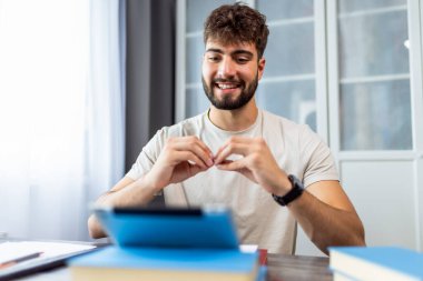 Genç bir öğrenci masada kitaplarla oturuyor, işaret dilini öğreniyor ve tabletinde el hareketleri yapıyor, iletişim becerilerini geliştirmeye odaklanıyor..