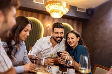 Genç çift modern kafede arkadaşlarıyla telefon ekranını paylaşıyor, sosyal anın ve gündelik etkileşimin tadını çıkarıyor..