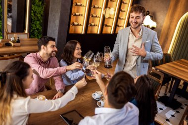 Bir grup genç arkadaş modern kafede kadeh kaldırırken bir adam ayakta duruyor, neşeli sosyal an..