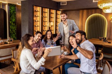 Modern kafede oturan bir grup genç arkadaş video görüşmesi için tablet kullanıyorlar, uzaktan biriyle bağlantı kuruyorlar..