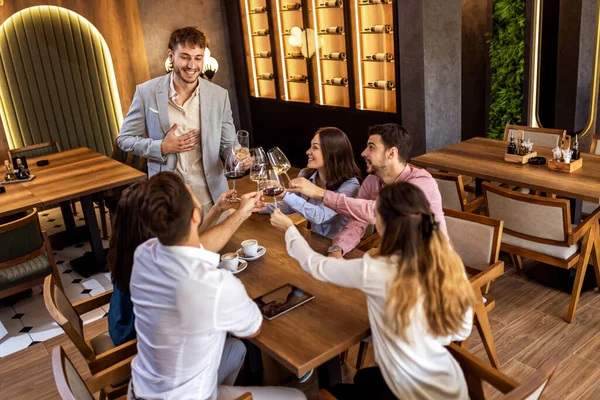 Bir grup genç arkadaş modern kafede kadeh kaldırırken bir adam ayakta duruyor, neşeli sosyal an..