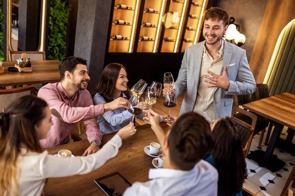 Bir grup genç arkadaş modern kafede kadeh kaldırırken bir adam ayakta duruyor, neşeli sosyal an..