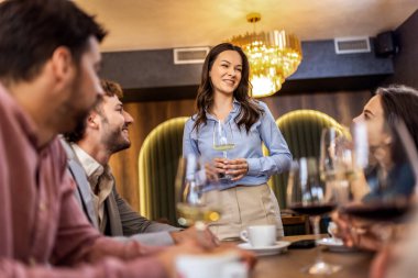 Bir grup arkadaş oturup dinlerken, modern kafede dikilen ve konuşan genç bir kadın..