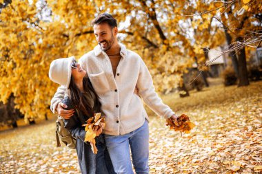 Gülümseyen genç çift sonbahar parkında yürüyor, renkli yaprakları bir arada tutuyorlar ve romantik sonbahar gününün tadını çıkarıyorlar..