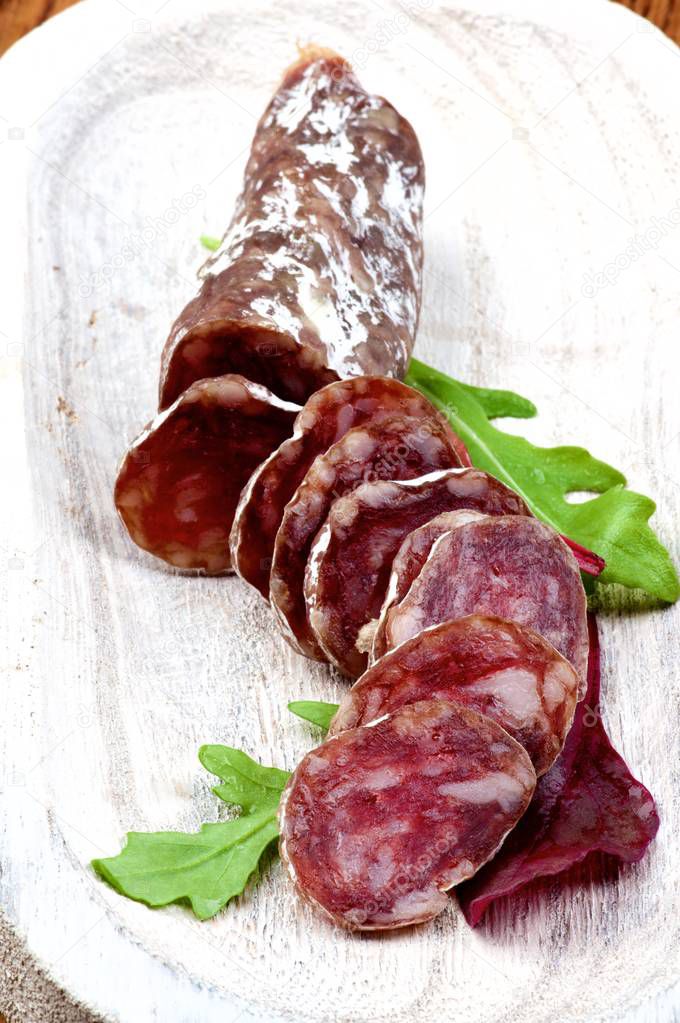 Slices and Half of Delicious Jerked Sausage Salchichon with Greens in White Wooden Plate closeup