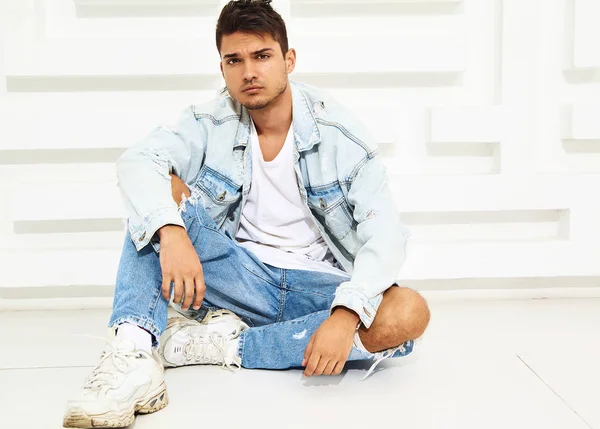 Portrait of handsome young model man dressed in jeans clothes sitting near white textured wall