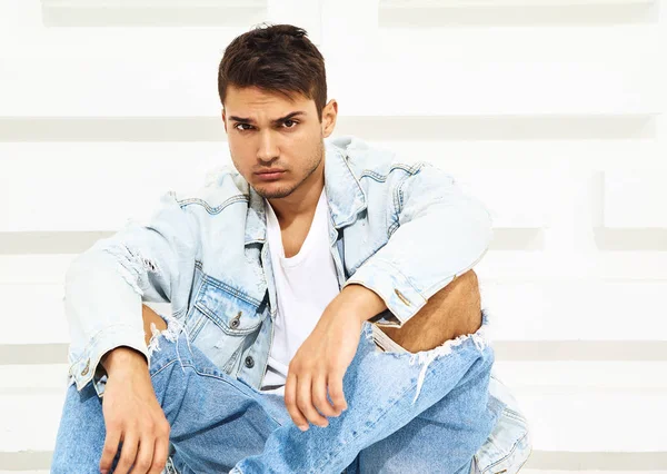 ホワイト テクスチャ壁のそばに座ってジーンズ服を着てハンサムな若いモデルの男のポートレート — ストック写真