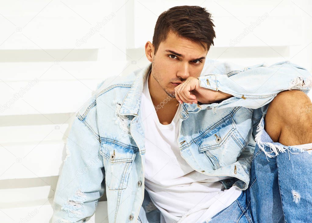 Portrait of handsome young model man dressed in jeans clothes sitting near white textured wall