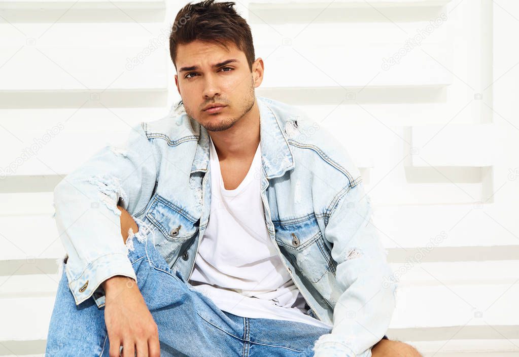 Portrait of handsome young model man dressed in jeans clothes sitting near white textured wall