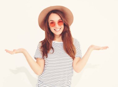 Karşılaştırma kavramı. Genç esmer kadın rahat hipster giysiler ve bir ürün, benzer seçimi için düz iki elle görüntülemek kahverengi şapka üzerine beyaz izole. İçinde güneş gözlüğü