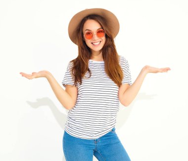 Karşılaştırma kavramı. Genç esmer kadın rahat hipster giysiler ve bir ürün, benzer seçimi için düz iki elle görüntülemek kahverengi şapka üzerine beyaz izole. İçinde güneş gözlüğü