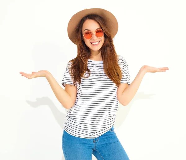 Karşılaştırma kavramı. Genç esmer kadın rahat hipster giysiler ve bir ürün, benzer seçimi için düz iki elle görüntülemek kahverengi şapka üzerine beyaz izole. İçinde güneş gözlüğü