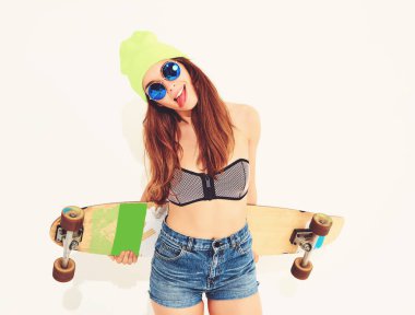 Genç şık gülümseyen kız modeli rahat yaz mayo giysiler ve sarı beanie longboard resepsiyon ile poz portresi. Dilini gösterilen beyaz üzerinde izole