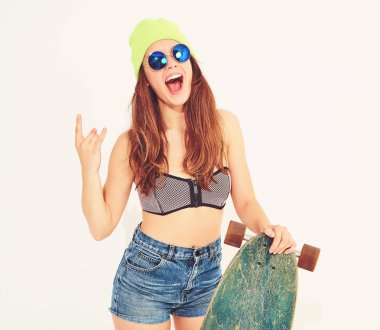 Genç şık gülümseyen kız modeli rahat yaz mayo giysiler ve sarı beanie rock'n roll işaret gösteren longboard resepsiyon ile poz. Beyaz izole