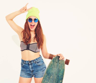 Genç şık gülümseyen kız modeli rahat yaz mayo giysiler ve sarı beanie rock'n roll işaret gösteren longboard resepsiyon ile poz. Beyaz izole