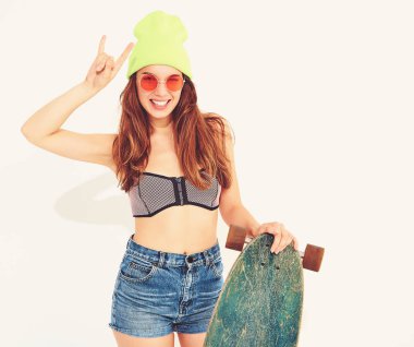 Genç şık gülümseyen kız modeli rahat yaz mayo giysiler ve sarı beanie rock'n roll işaret gösteren longboard resepsiyon ile poz. Beyaz izole