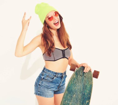 Genç şık gülümseyen kız modeli rahat yaz mayo giysiler ve sarı beanie rock'n roll işaret gösteren longboard resepsiyon ile poz. Beyaz izole