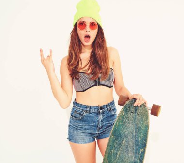 Genç şık gülümseyen kız modeli rahat yaz mayo giysiler ve sarı beanie rock'n roll işaret gösteren longboard resepsiyon ile poz. Beyaz izole