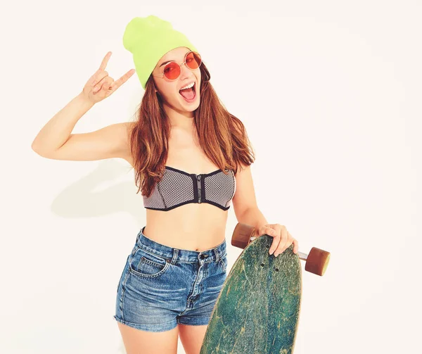 Genç şık gülümseyen kız modeli rahat yaz mayo giysiler ve sarı beanie rock'n roll işaret gösteren longboard resepsiyon ile poz. Beyaz izole