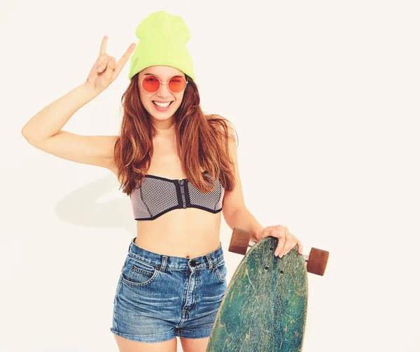 Genç şık gülümseyen kız modeli rahat yaz mayo giysiler ve sarı beanie rock'n roll işaret gösteren longboard resepsiyon ile poz. Beyaz izole
