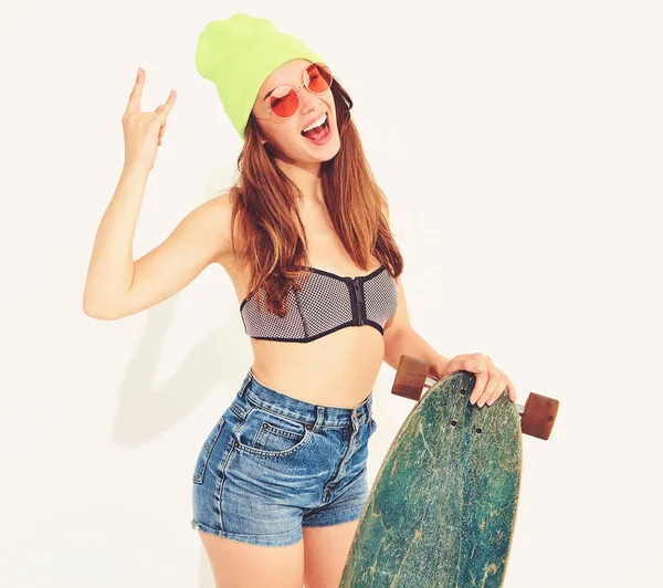 Genç şık gülümseyen kız modeli rahat yaz mayo giysiler ve sarı beanie rock'n roll işaret gösteren longboard resepsiyon ile poz. Beyaz izole