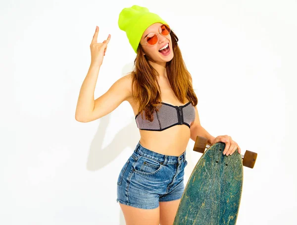 Genç şık gülümseyen kız modeli rahat yaz mayo giysiler ve sarı beanie rock'n roll işaret gösteren longboard resepsiyon ile poz. Beyaz izole
