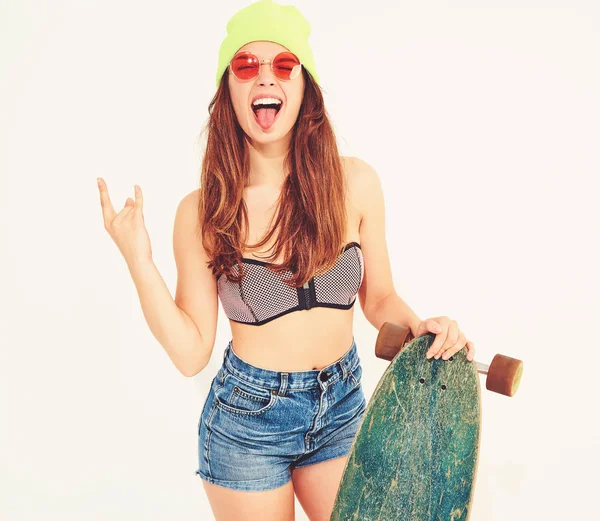 Genç şık gülümseyen kız modeli rahat yaz mayo giysiler ve sarı beanie rock'n roll işaret gösteren longboard resepsiyon ile poz. Beyaz izole
