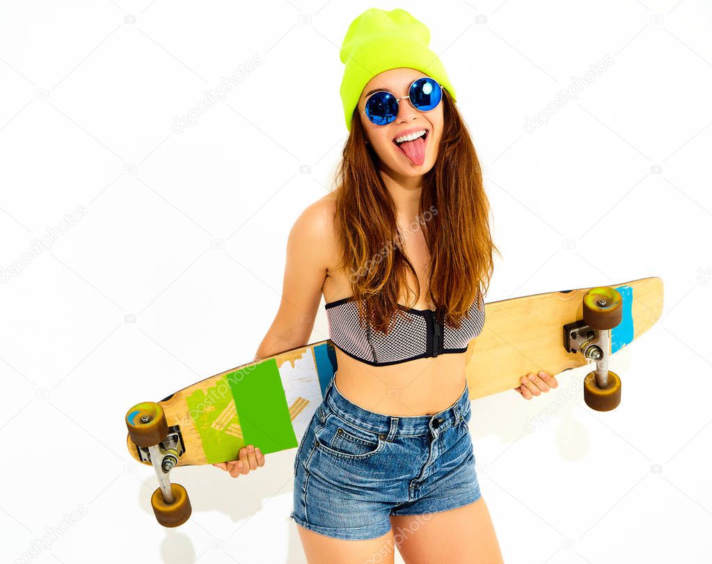 Portrait of young stylish smiling girl model in casual summer swimwear clothes and yellow beanie posing with longboard desk. Isolated on white, showing her tongue