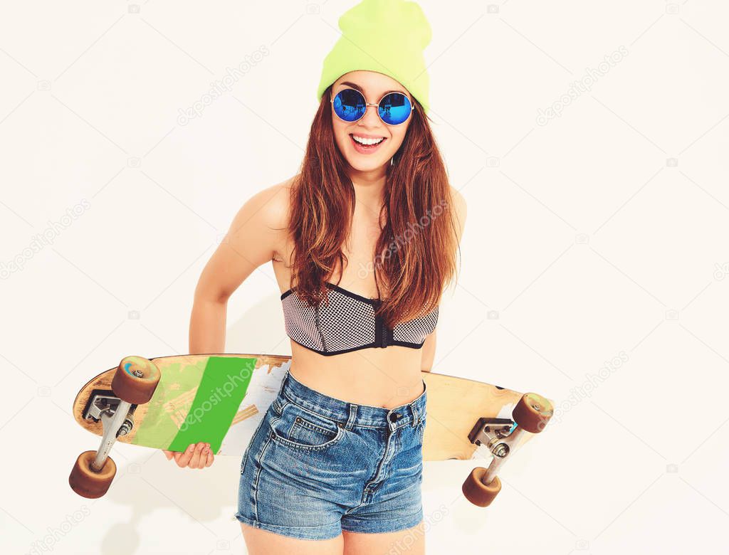 Portrait of young stylish smiling girl model in casual summer swimwear clothes and yellow beanie posing with longboard desk. Isolated on white