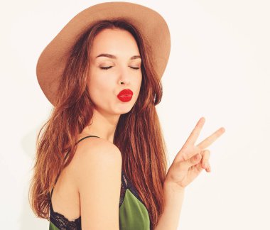 Genç şık kız modeli rahat yaz yeşil giysiler ve kırmızı dudaklar ile kahverengi şapka. Barış işareti gösterilen ve öpücük vererek