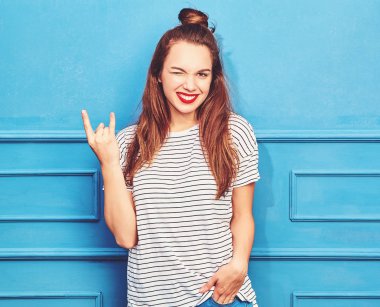 Gündelik yaz kıyafetleri kırmızı dudaklar, mavi duvara yakın poz ile genç şık kız modeli. Göz kırpıyor ve rock'n roll işaret gösteren