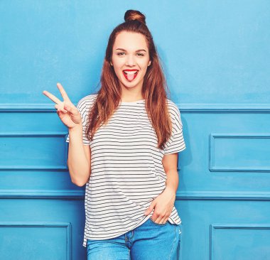 Gündelik yaz kıyafetleri kırmızı dudaklar, mavi duvara yakın poz ile genç şık kız modeli. Onun dilini ve barış işareti gösteren