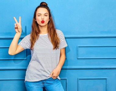 Gündelik yaz kıyafetleri kırmızı dudaklar, mavi duvara yakın poz ile genç şık kız modeli. Öpücük ve barış işareti veren