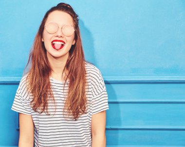 Genç şık kız modeli güneş gözlüğü mavi duvarının yakınında poz kırmızı dudaklar ile rahat yaz giysileri. Dilini gösterilen