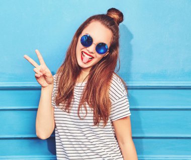 Gündelik yaz kıyafetleri kırmızı dudaklar, mavi duvara yakın poz ile genç şık kız modeli. Barış işareti ve dilini gösteren