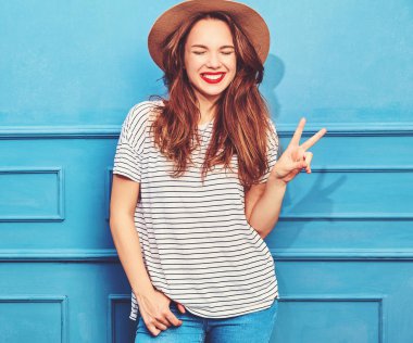 Gündelik yaz kıyafetleri ve kahverengi şapka kırmızı dudaklar, mavi duvara yakın poz ile genç şık kız modeli. Barış işareti ve dilini gösteren