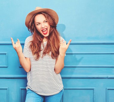 Gündelik yaz kıyafetleri ve kahverengi şapka kırmızı dudaklar, mavi duvara yakın poz ile genç şık kız modeli. Rock'n roll işaret gösteren