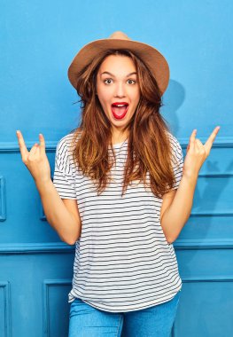 Gündelik yaz kıyafetleri ve kahverengi şapka kırmızı dudaklar, mavi duvara yakın poz ile genç şık kız modeli. Rock'n roll işaret gösteren