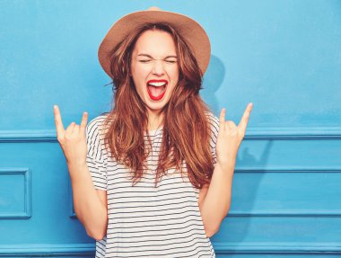 Gündelik yaz kıyafetleri ve kahverengi şapka kırmızı dudaklar, mavi duvara yakın poz ile genç şık kız modeli. Rock'n roll işaret gösteren