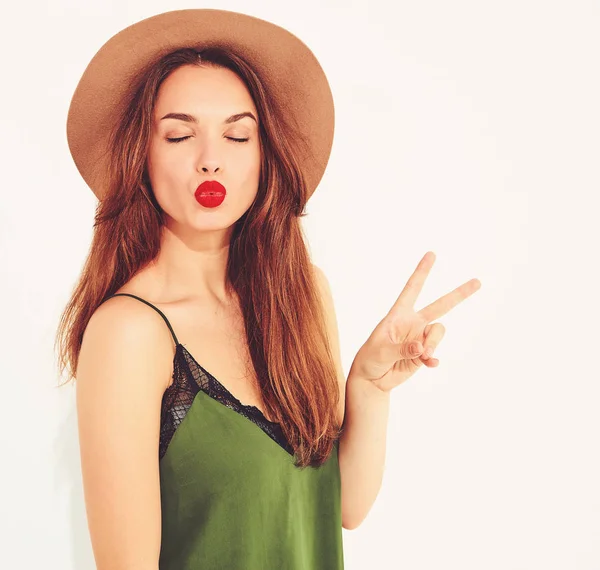 Genç şık kız modeli rahat yaz yeşil giysiler ve kırmızı dudaklar ile kahverengi şapka. Barış işareti gösterilen ve öpücük vererek