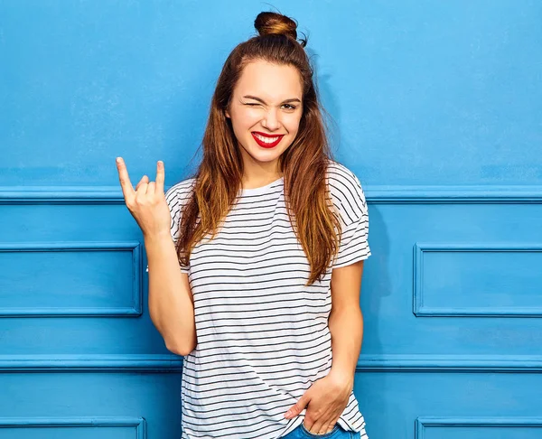 Gündelik yaz kıyafetleri kırmızı dudaklar, mavi duvara yakın poz ile genç şık kız modeli. Göz kırpıyor ve rock'n roll işaret gösteren