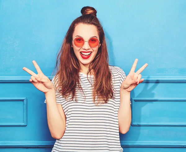 Gündelik yaz kıyafetleri kırmızı dudaklar, mavi duvara yakın poz ile genç şık kız modeli. Barış işareti gösterilen