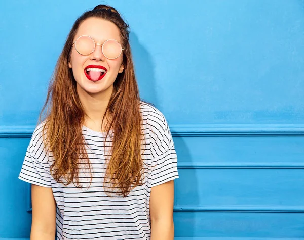 Genç şık kız modeli güneş gözlüğü mavi duvarının yakınında poz kırmızı dudaklar ile rahat yaz giysileri. Dilini gösterilen