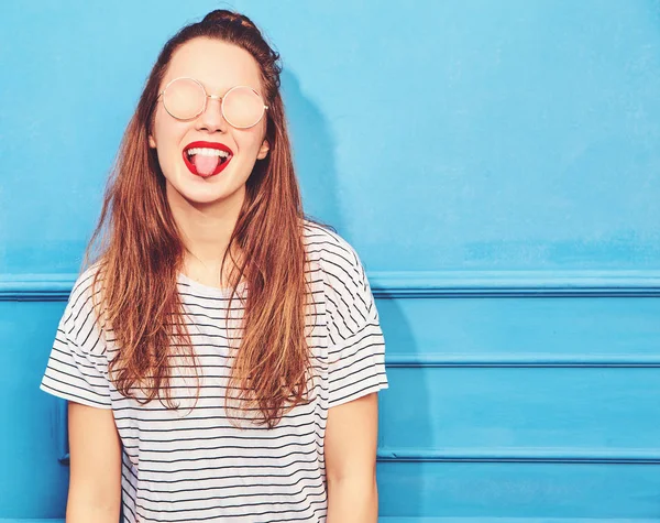 Genç şık kız modeli güneş gözlüğü mavi duvarının yakınında poz kırmızı dudaklar ile rahat yaz giysileri. Dilini gösterilen