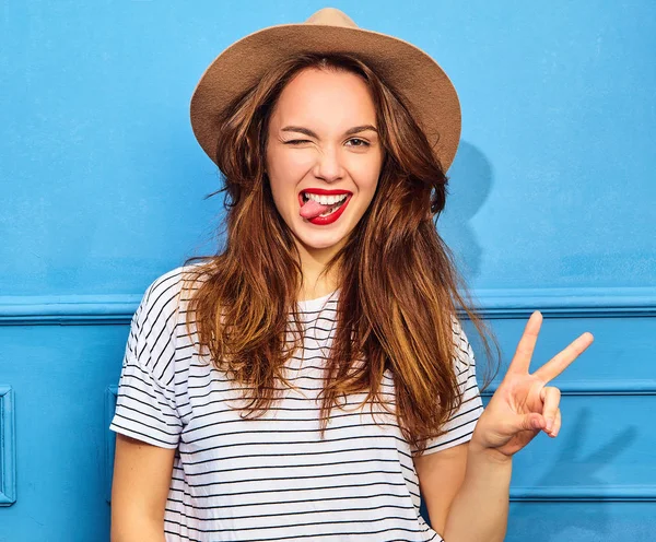 Gündelik yaz kıyafetleri ve kahverengi şapka kırmızı dudaklar, mavi duvara yakın poz ile genç şık kız modeli. Göz kırpıyor ve barış işareti gösterilen